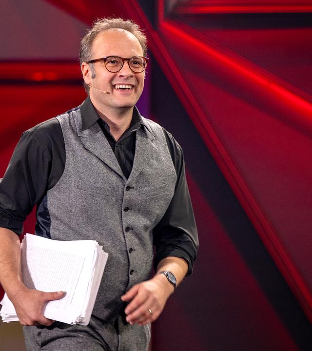 René Sydow ist in der Mitte des Bildes zu sehen. Er trägt einen Stapel Papier unter dem rechten Arm. Der Hintergrund ist rot beleuchtet.