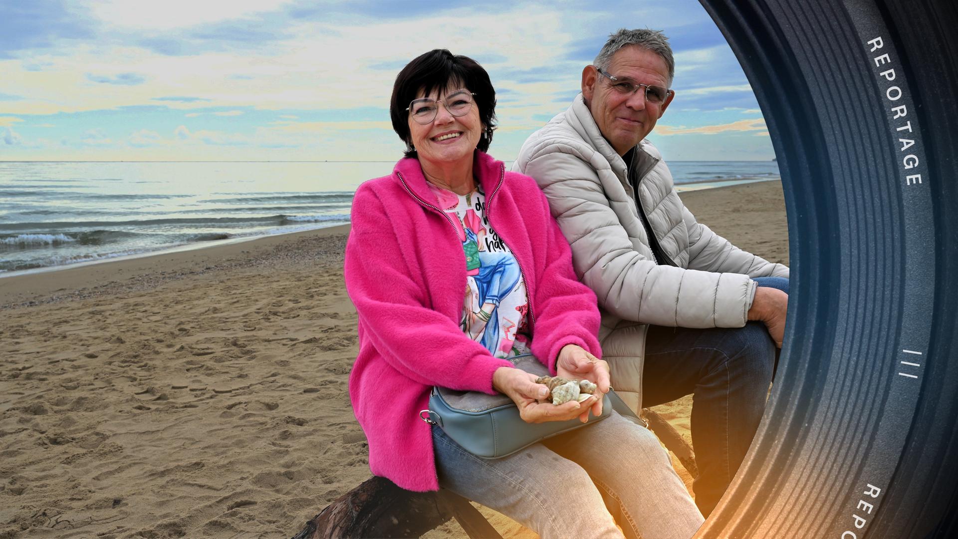 Zwei Senioren sitzen an einem Strand und lächeln in die Kamera. Im Hintergrund ist das Meer zu sehen.