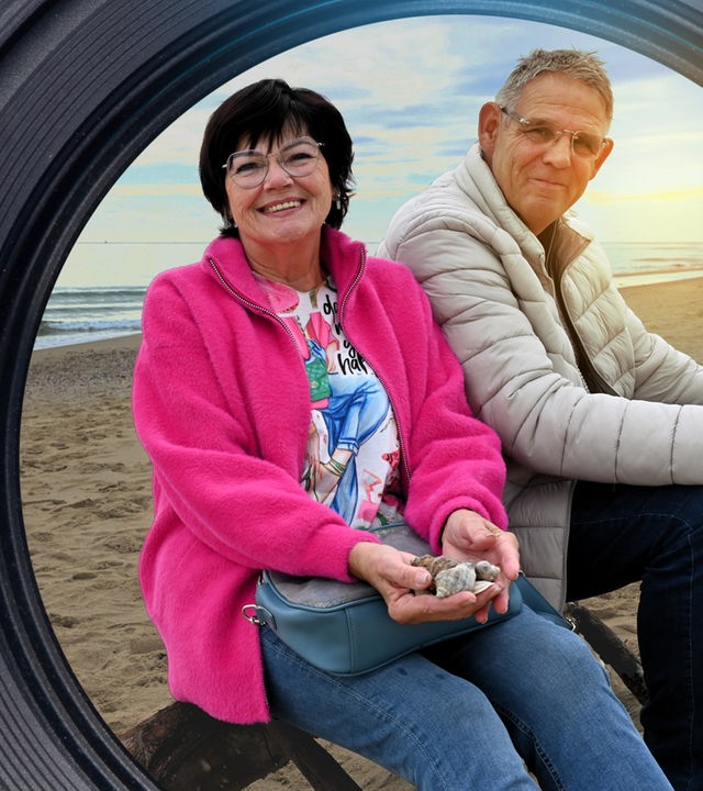 Zwei Senioren sitzen an einem Strand und lächeln in die Kamera. Im Hintergrund ist das Meer zu sehen.