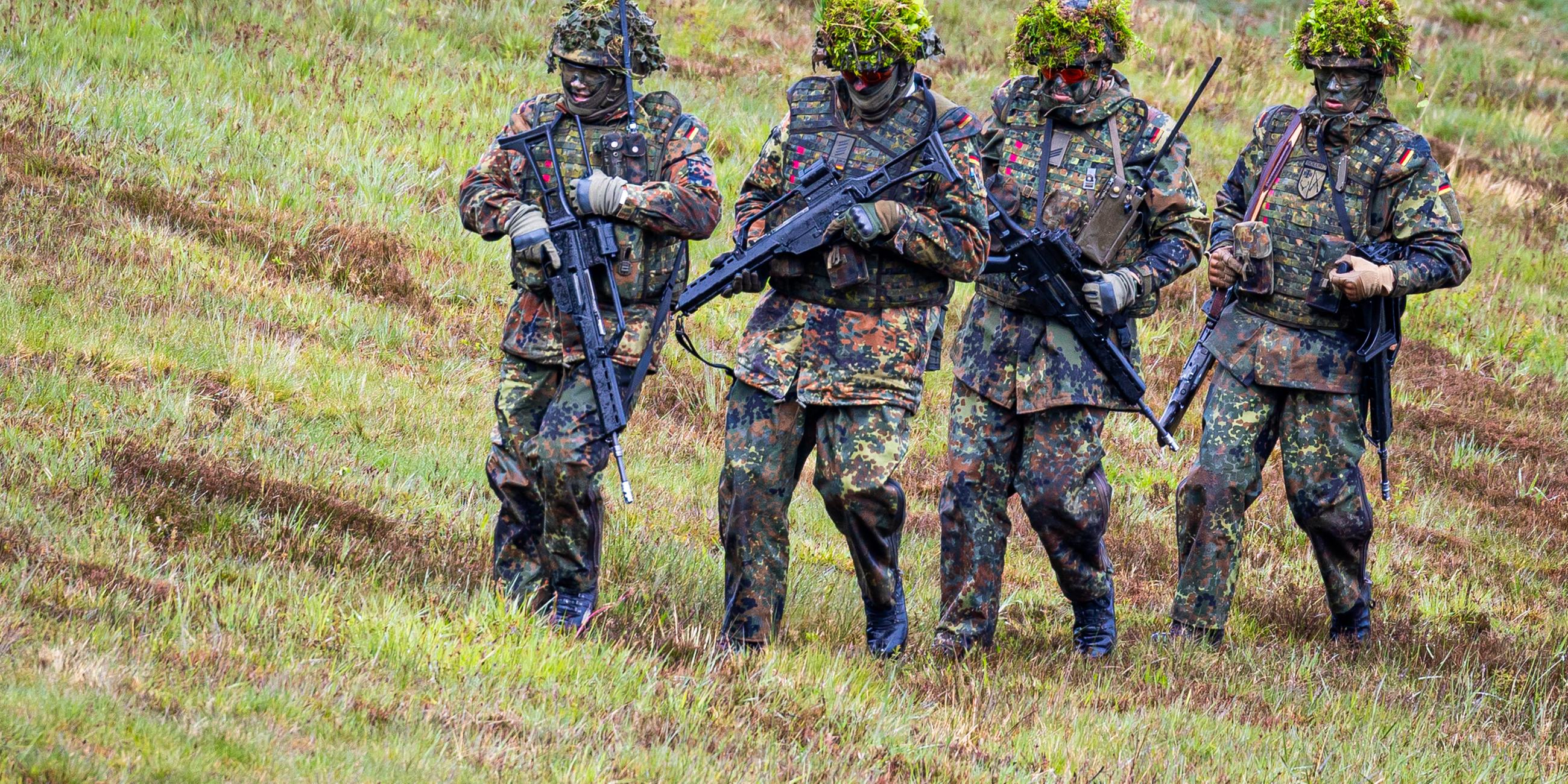 Vier vermumte Personen in militärischem Camouflage mit Zweigen auf den Helmen und Waffen in den Händen stehen etwas ungelenk nebeneinander auf einer Wiese.