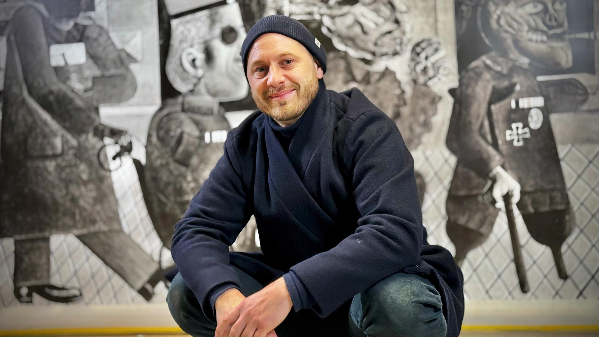 Ein Mann sitzt in Hockstellung vor einem Gemälde und lächelt in die Kamera. Er trägt eine dunkelblaue Mütze und Jacke und eine Jeans. Das Gemälde hinter ihm ist ein großformatiges schwarz-weiß Kunstwerk.
