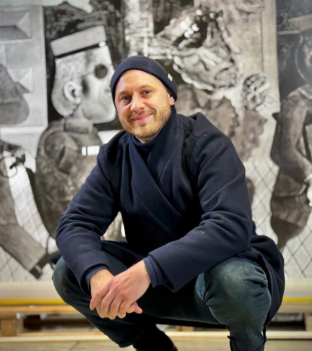 Ein Mann sitzt in Hockstellung vor einem Gemälde und lächelt in die Kamera. Er trägt eine dunkelblaue Mütze und Jacke und eine Jeans. Das Gemälde hinter ihm ist ein großformatiges schwarz-weiß Kunstwerk.