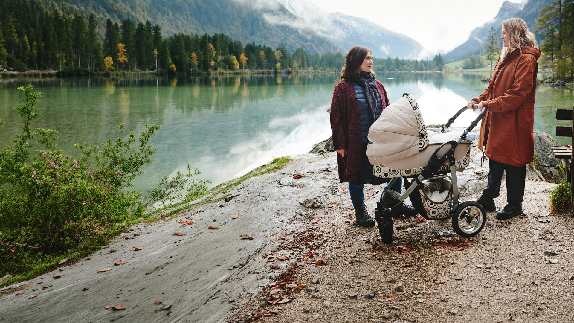 "Lena Lorenz - Retterbaby": An einem See in Berglandschaft stehen Eva (Eva Mattes) und Lena Lorenz (Judith Hoersch) mit Kinderwagen und schauen sich an.