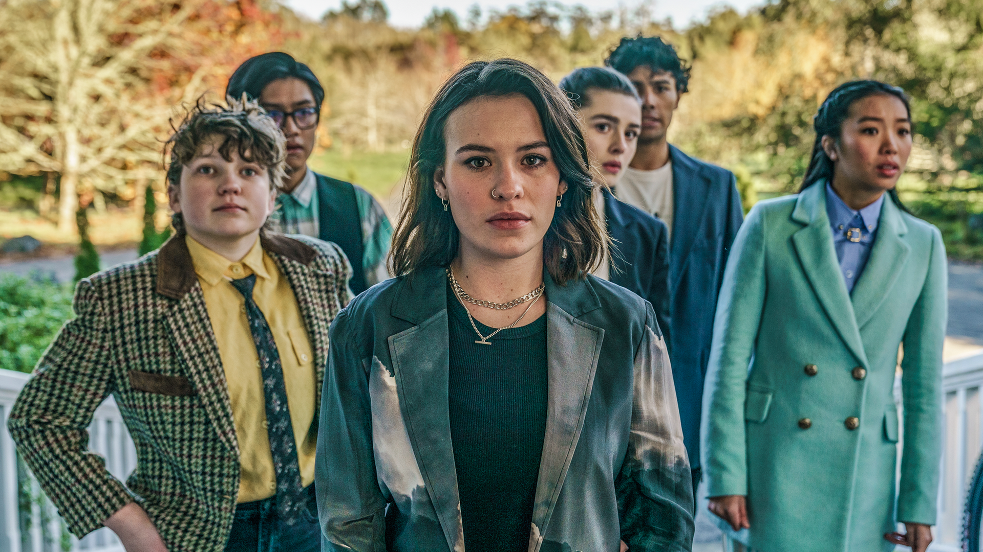 Issie, Stella, Natascha, Caroline, Caleb und Dan stehen als Gruppe mit angespannt fragenden Gesichtern auf einem Treppenaufgang an dem seitlich weiße Holzgeländer hochführen. Im Hintergrund sieht man Bäume.