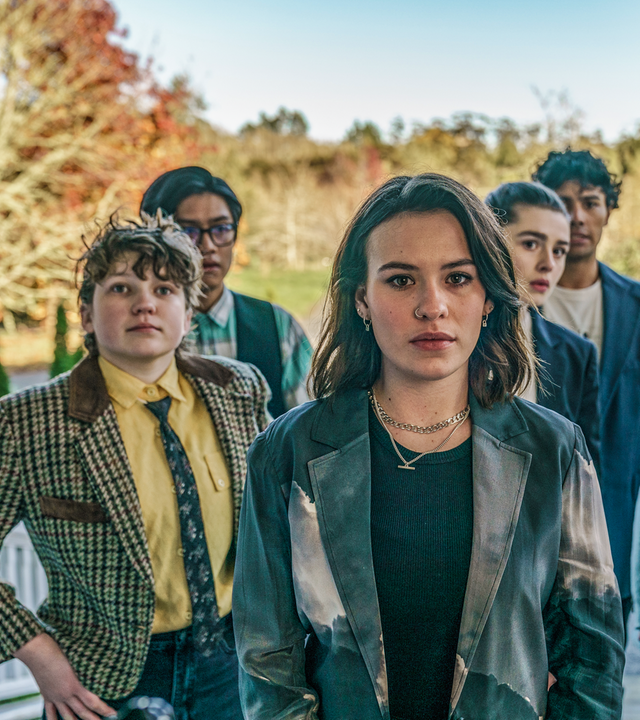 Issie, Stella, Natascha, Caroline, Caleb und Dan stehen als Gruppe mit angespannt fragenden Gesichtern auf einem Treppenaufgang an dem seitlich weiße Holzgeländer hochführen. Im Hintergrund sieht man Bäume.