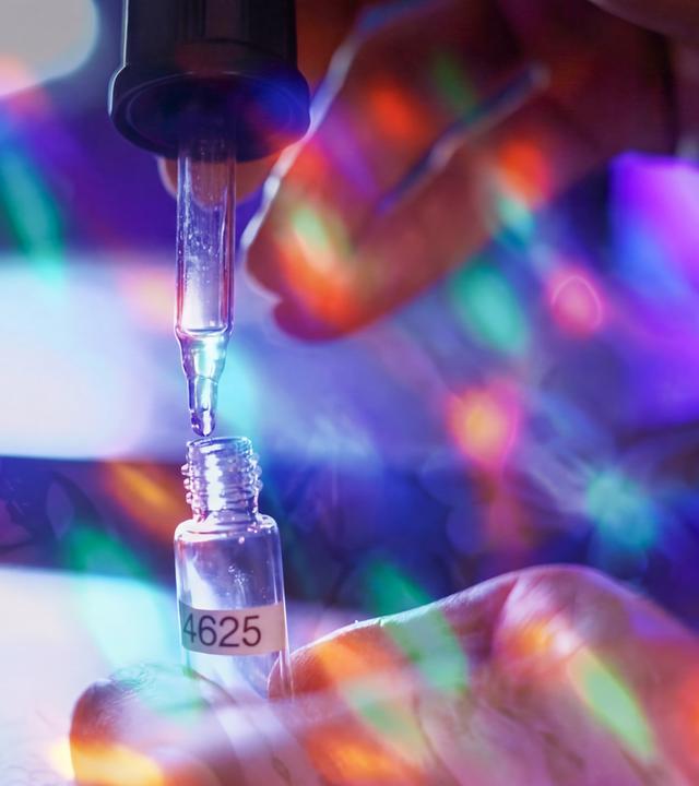 Nahaufnahme einer Pipette, die eine klare Flüssigkeit in ein kleines beschriftetes Fläschchen tropft; eine Hand hält das Gefäß, bunte Lichtreflexe überlagern die Laborszene.