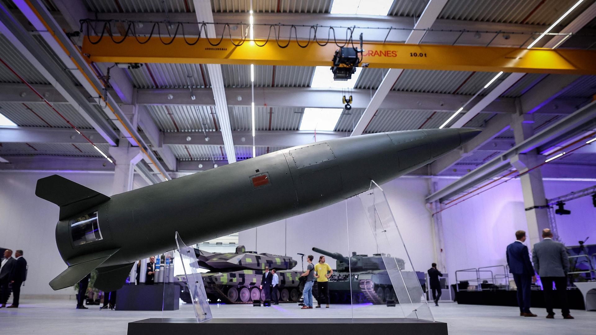 In einer großen Werkshalle ist im Vordergrund eine auf durchsichtigen Stützen aufgestellte Rakete zu sehen. Im Hintergrund stehen Menschen um zwei Panzer.