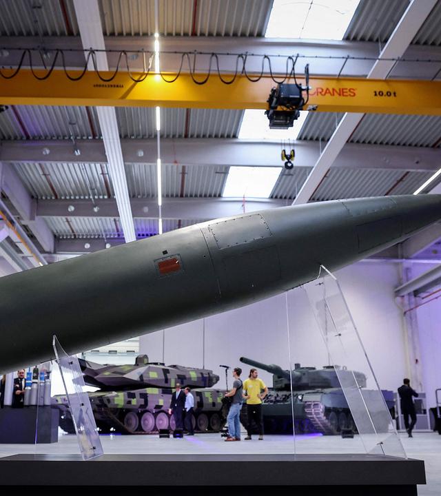 In einer großen Werkshalle ist im Vordergrund eine auf durchsichtigen Stützen aufgestellte Rakete zu sehen. Im Hintergrund stehen Menschen um zwei Panzer.