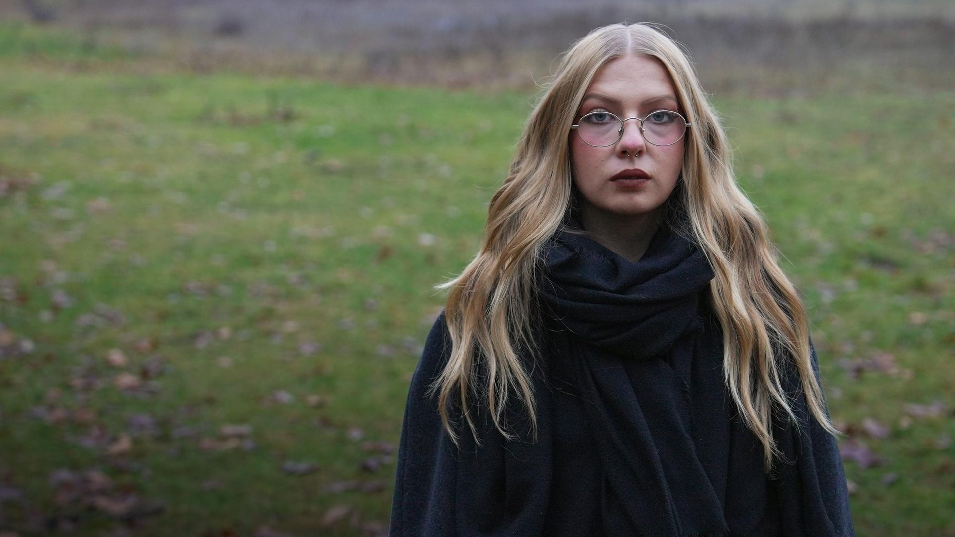 Ricarda  steht im auf einer Wiese und schaut starr in die Kamera. 