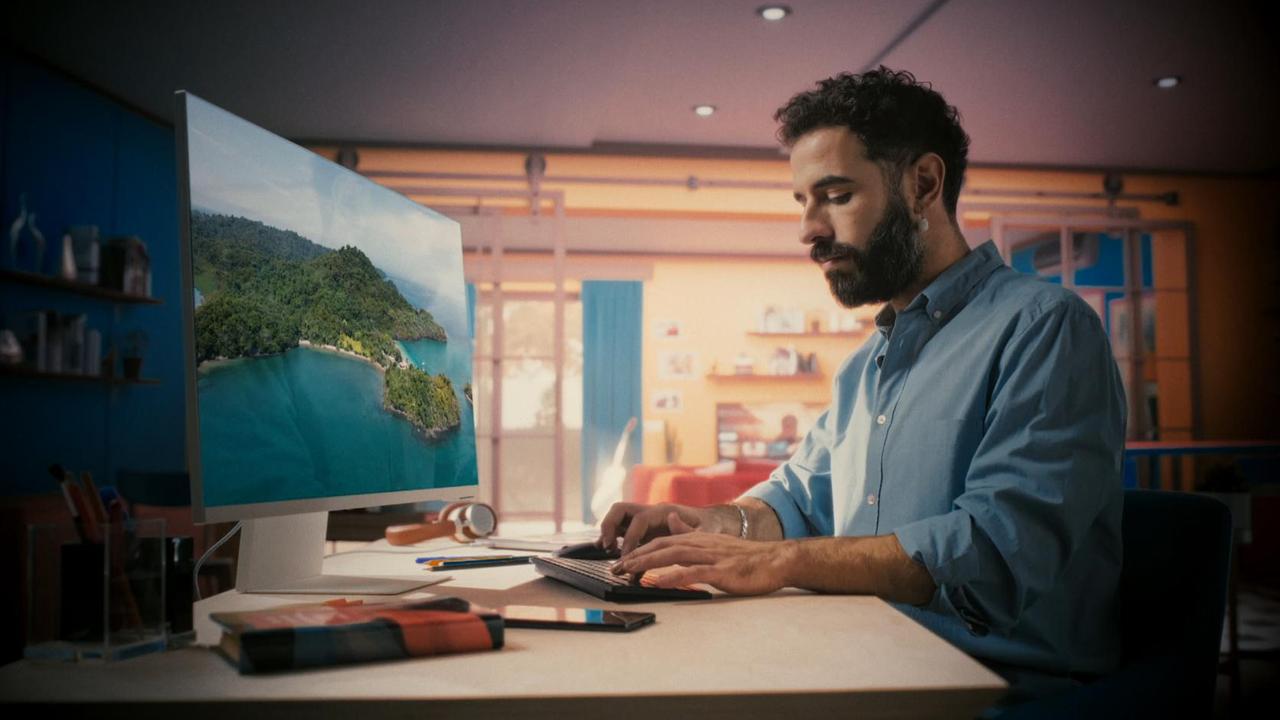 Szene aus "Das große Spiel" von Richard Powers: Seitlicher Blick auf einen Mann, der an einem Schreibtisch mit Bildschirm und Tastatur sitzt. Seine Hände liegen auf der Tastatur. Sein Blick geht auf die Tastur und seine schreibenden Hände. Auf dem Bildschirm ist eine bewaldete Insel mit einer Bucht und einem Sandtstrand zu sehen. Auf dem Schreibtisch befinden sich noch ein Kopfhörer, Kalender und Stifte. Im Hintergrund des Zimmers befindet sich eine Balkontür. An der Wand rechts daneben befinden sich Regalbretter mit Büchern. Der Hindergrund ist weichgezeichnet.