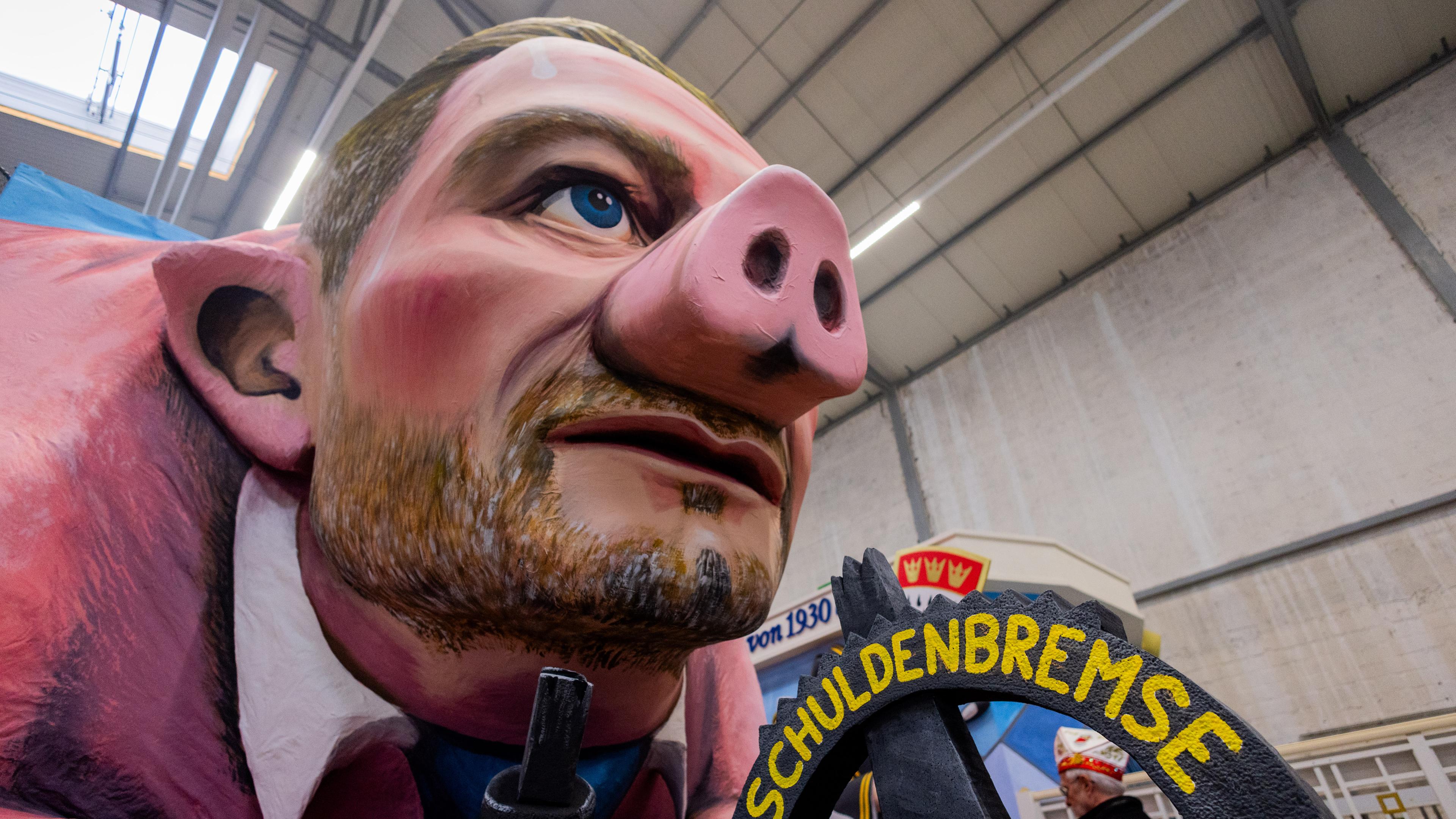 Ein Motivwagen, der Bundesfinanzminister Christian Lindner als Sparschwein mit der Schuldenbremse zeigt, steht beim Richtfest für den Kölner Rosenmontagszug im Karnevalsmuseum.