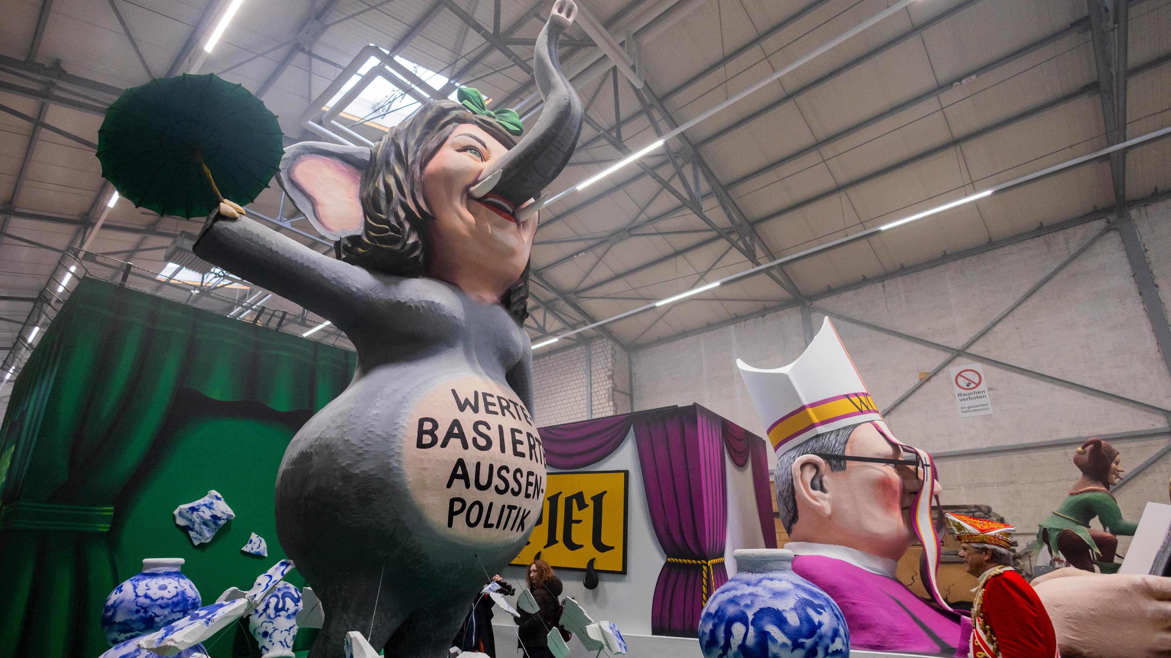 Zwei Motivwagen, die Bundesaußenministerin Annalena Baerbock als Elefant im Porzellanladen zeigen, stehen beim Richtfest für den Kölner Rosenmontagszug im Karnevalsmuseum.