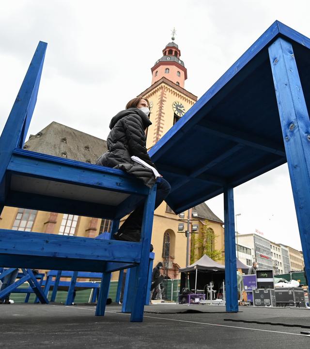 Ein Riesen-Tisch und ein Riesen-Stuhl vor einer Kirche, eine Frau mit Mundschutz sitzt auf dem Stuhl.