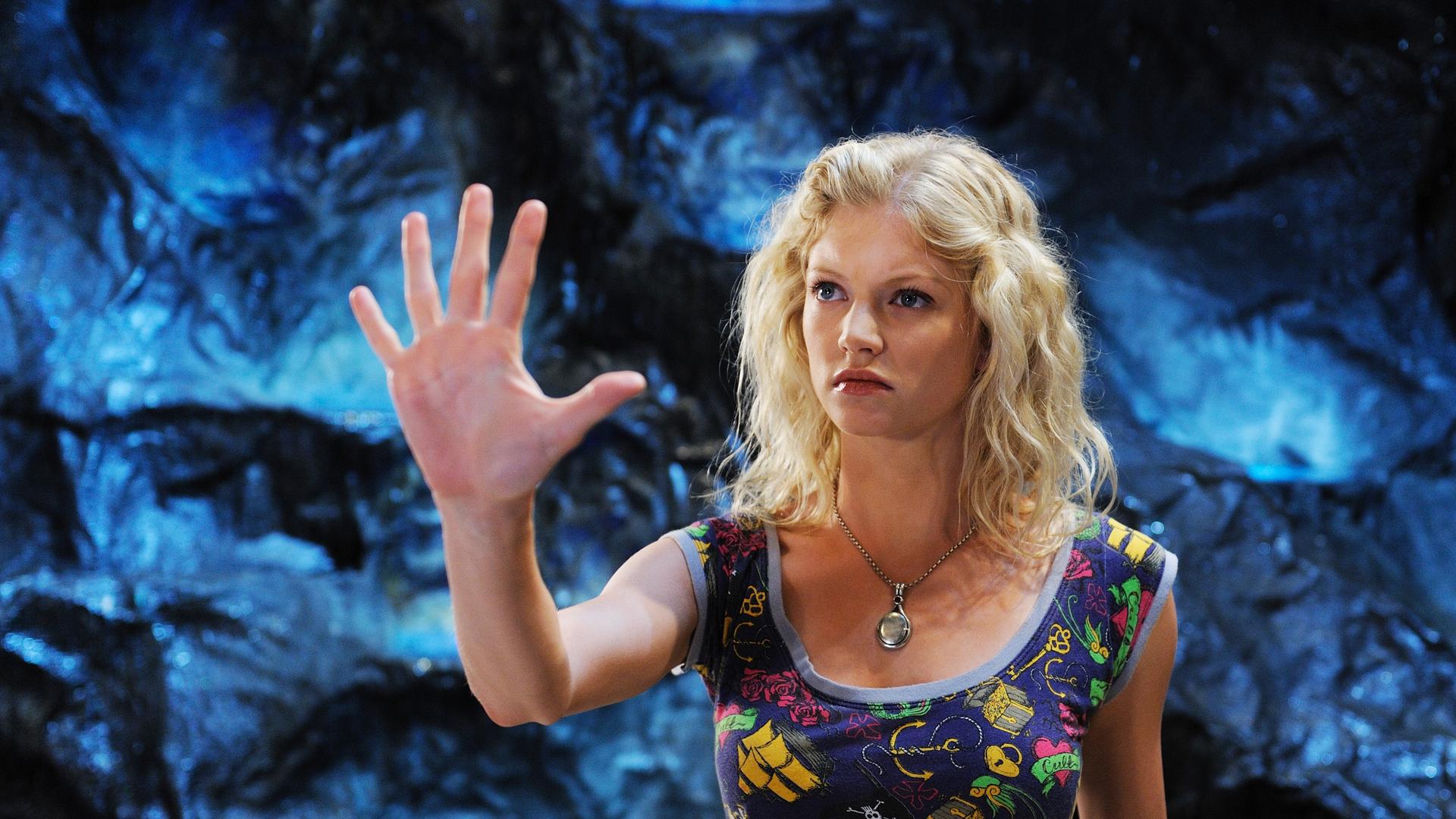 Rikki steht in der Grotte auf Mako Island vor einer blau glänzenden Felswand. Sie hält konzentriert die erhobene Hand vor sich, ihre Finger sind weit auseinandergespreizt.
