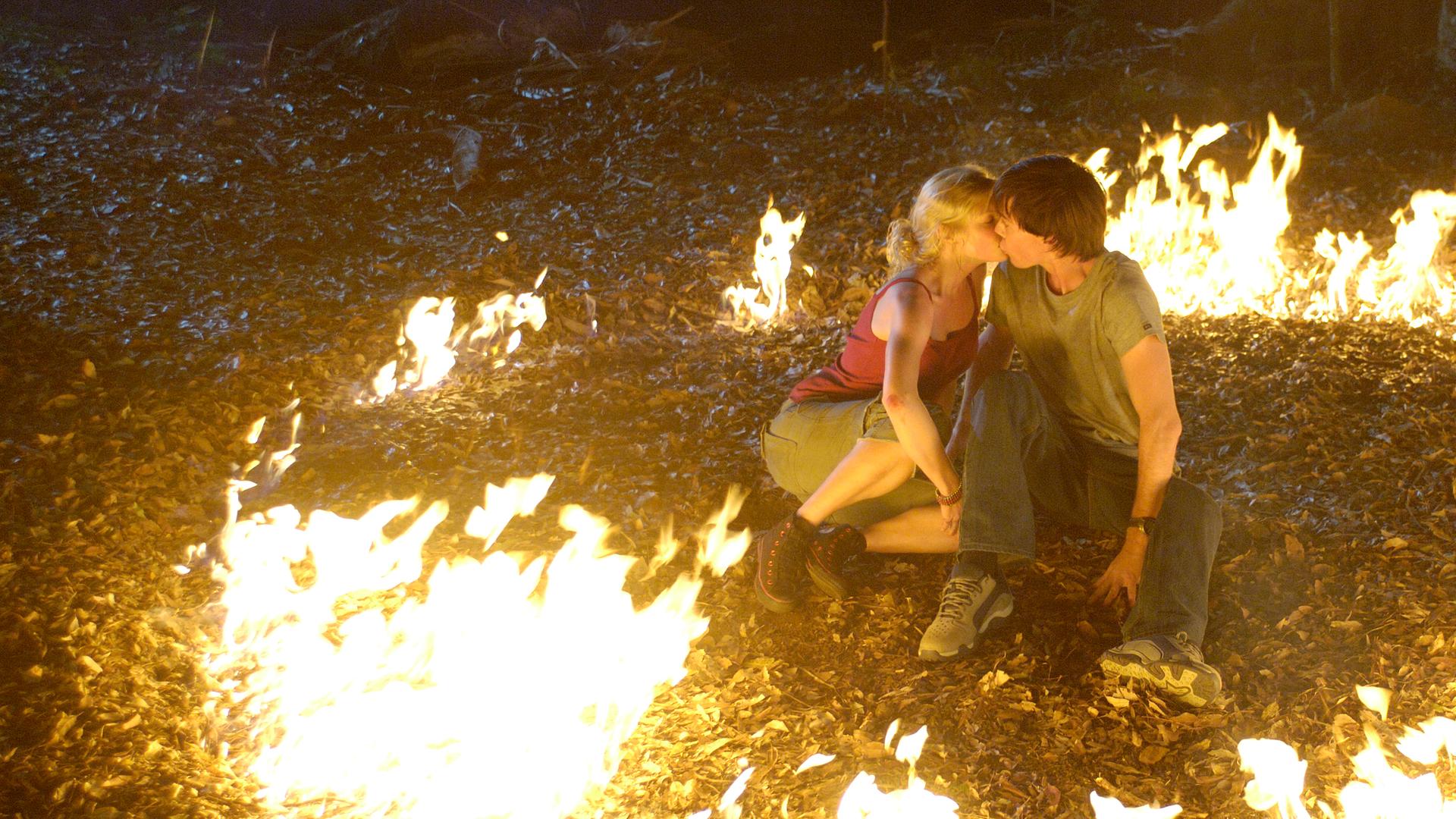 Rikki und Zane sitzen im Wald auf dem Boden und küssen sich. Um sie herum ein Kreis aus Flammen.