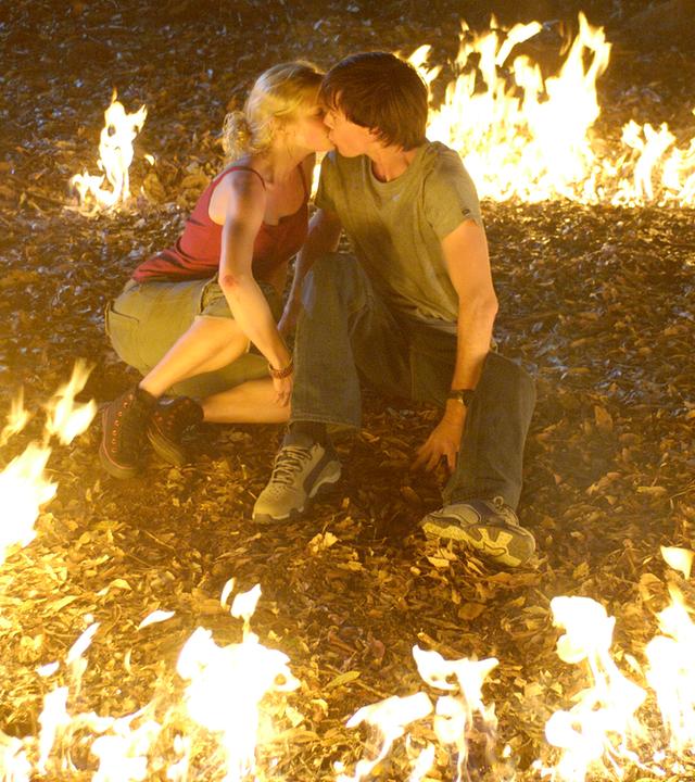 Rikki und Zane sitzen im Wald auf dem Boden und küssen sich. Um sie herum ein Kreis aus Flammen.