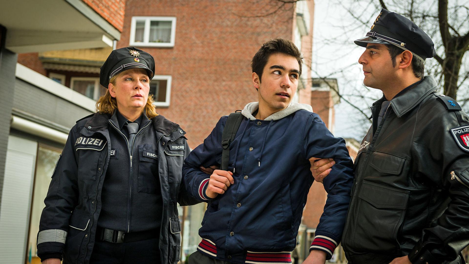 "Notruf Hafenkante - Ringo, Kim und Sidney": Ringo (Tan Julius Ipekkaya) steht zwischen den Polizisten Claudia Fischer (Janette Rauch) und Tarik Coban (Serhat Cokgezen), die ihn an den Armen festhalten. Tarik sieht den Jungen böse an.