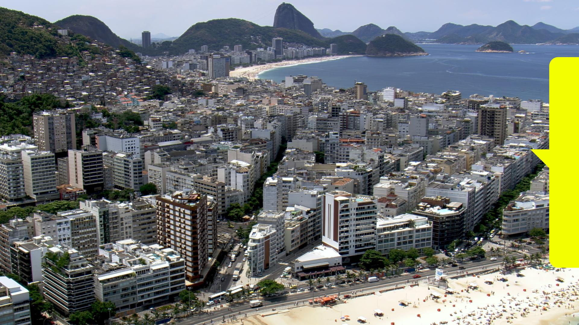 Blick von schräg oben: Ein breiter, mit vielen Sonnenbadenden besetzter Strand liegt vor einer Stadt und bewaldeten Hügeln. Im Hintergrund liegt eine weitere Bucht.