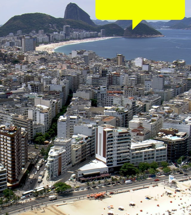 Blick von schräg oben: Ein breiter, mit vielen Sonnenbadenden besetzter Strand liegt vor einer Stadt und bewaldeten Hügeln. Im Hintergrund liegt eine weitere Bucht.