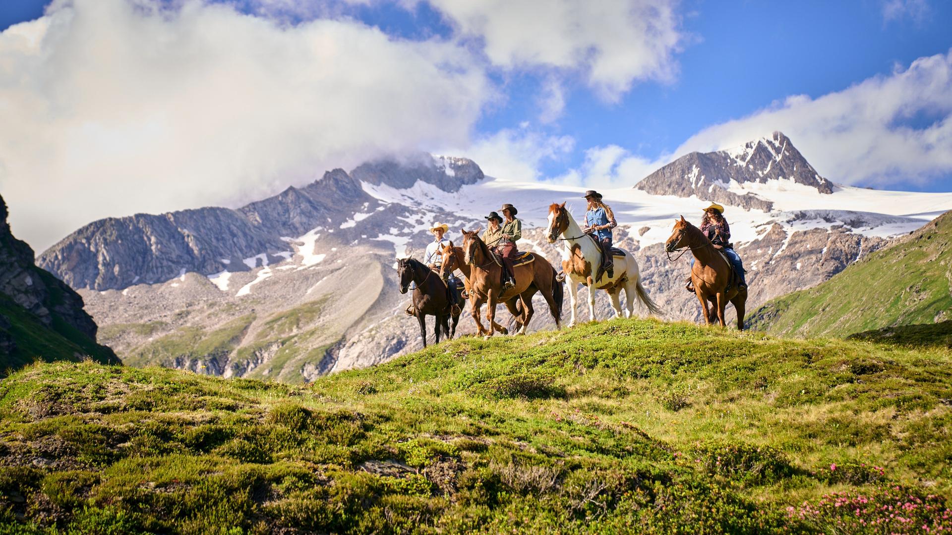 Das Bild zeigt eine Reitergruppe, die auf Pferden steht und eine malerische alpine Landschaft in Osttirol betrachtet. Im Hintergrund erhebt sich ein beeindruckendes Gebirge mit schneebedeckten Gipfeln unter einem klaren blauen Himmel, der mit wenigen Wolken durchzogen ist. Die Wiese, auf der die Reiter stehen, ist mit grünen Pflanzen und vereinzelten blühenden Sträuchern bedeckt. Die Reiter tragen westernmäßig angelegte Kleidung, darunter Hüte und Hemden, und wirken entspannt und in die Umgebung vertieft. Die Szene vermittelt ein Gefühl von Naturverbundenheit und Abenteuer.