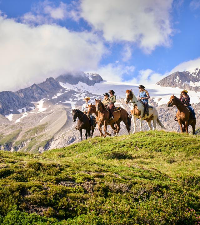 Das Bild zeigt eine Reitergruppe, die auf Pferden steht und eine malerische alpine Landschaft in Osttirol betrachtet. Im Hintergrund erhebt sich ein beeindruckendes Gebirge mit schneebedeckten Gipfeln unter einem klaren blauen Himmel, der mit wenigen Wolken durchzogen ist. Die Wiese, auf der die Reiter stehen, ist mit grünen Pflanzen und vereinzelten blühenden Sträuchern bedeckt. Die Reiter tragen westernmäßig angelegte Kleidung, darunter Hüte und Hemden, und wirken entspannt und in die Umgebung vertieft. Die Szene vermittelt ein Gefühl von Naturverbundenheit und Abenteuer.