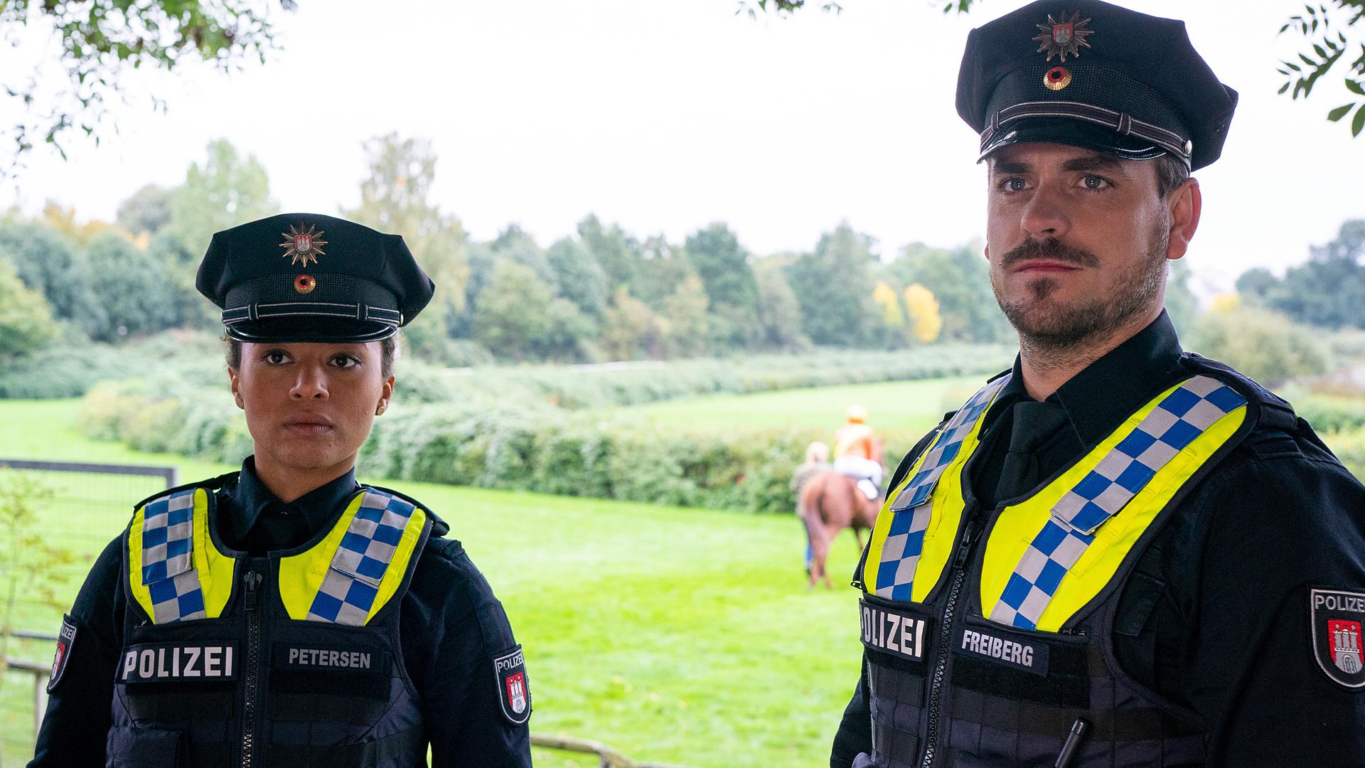 Die Polizistin Daisy Petersen (Aysha Joy Samuel) und ihr Kollege Kris Freiberg (Marc Barthel) stehen nebeneinander auf einem Gehweg. Hinter ihnen ist die Galopprennbahn zu sehen, sowie ein Jockey auf seinem Pferd. Beide beobachten etwas vor sich.