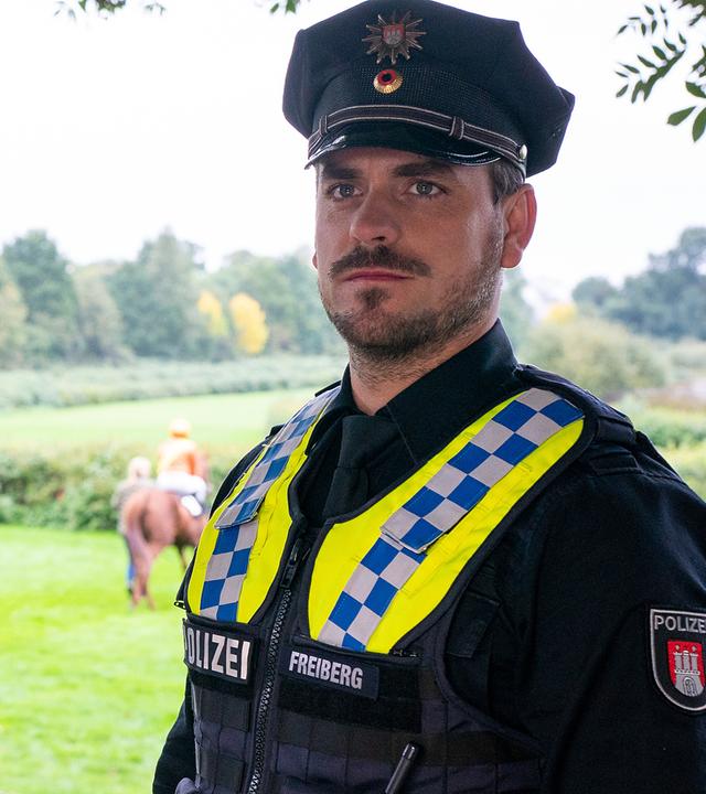 Die Polizistin Daisy Petersen (Aysha Joy Samuel) und ihr Kollege Kris Freiberg (Marc Barthel) stehen nebeneinander auf einem Gehweg. Hinter ihnen ist die Galopprennbahn zu sehen, sowie ein Jockey auf seinem Pferd. Beide beobachten etwas vor sich.