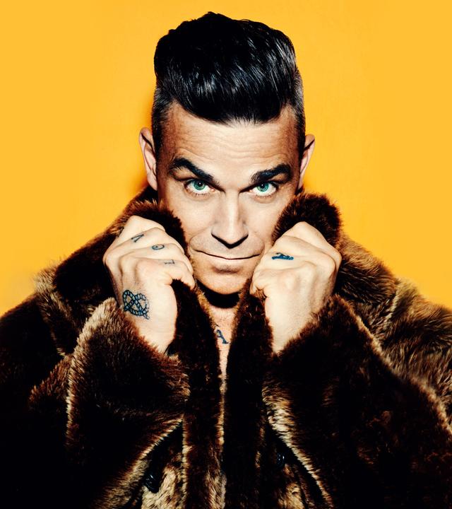 Robbie Williams vor einem gelben Hintergrund. Er trägt einen braunen Fellmantel und hält die Hände am Kragen.