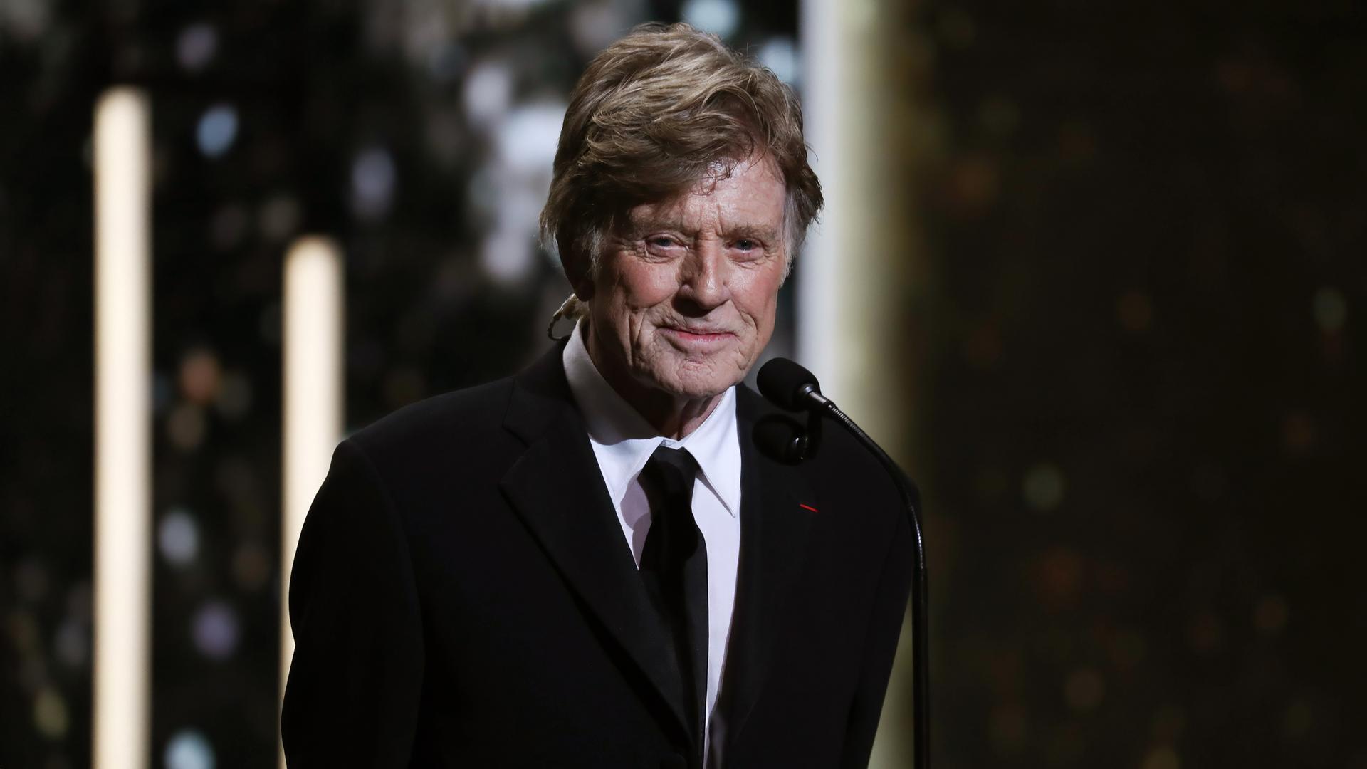 Porträtfoto eines alten Mannes im Smoking, der mit auf dem Rücken verschränkten Armen lächelnd vor einem Mikrofon steht.