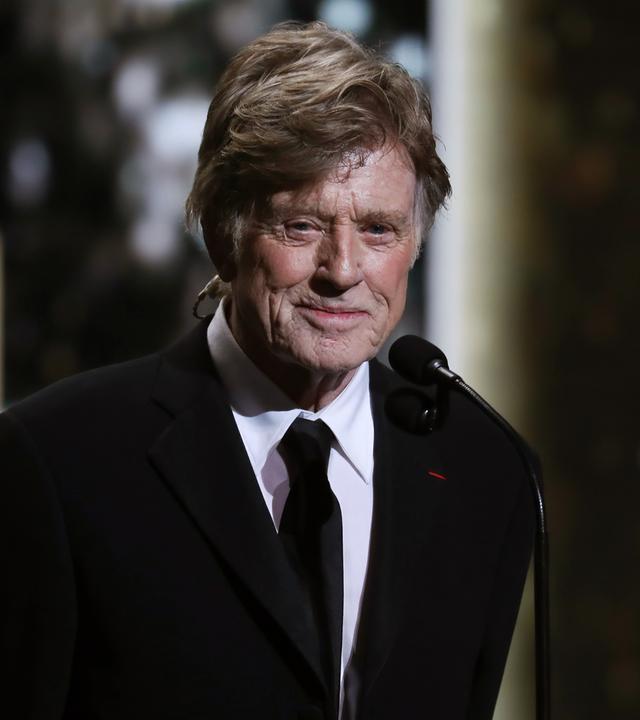 Porträtfoto eines alten Mannes im Smoking, der mit auf dem Rücken verschränkten Armen lächelnd vor einem Mikrofon steht.