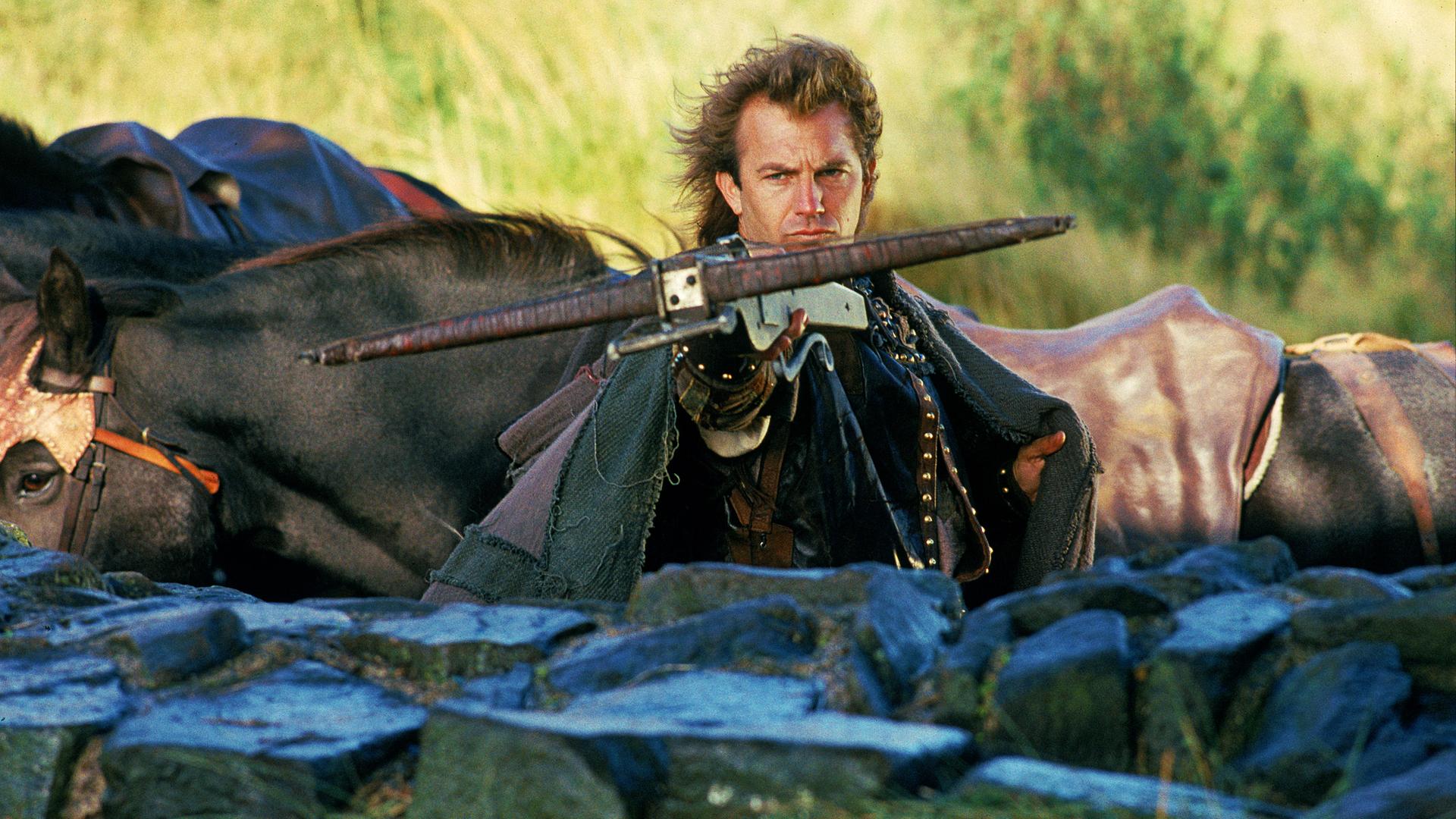 Robin Hood (Kevin Costner) steht hinter einer halbhohen Mauer und zielt mit einer Armbrust nach vorne. Im Hintergrund sind Pferde zu sehen.