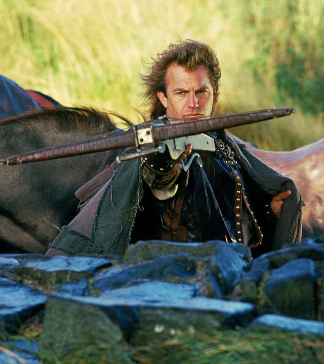 Robin Hood (Kevin Costner) steht hinter einer halbhohen Mauer und zielt mit einer Armbrust nach vorne. Im Hintergrund sind Pferde zu sehen.