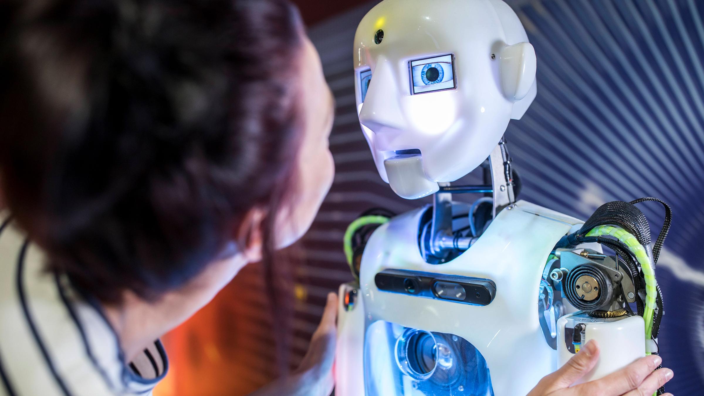 Frau beugt sich nach an einen Roboter in menschlicher Gestalt und betrachtet sein Gesicht.