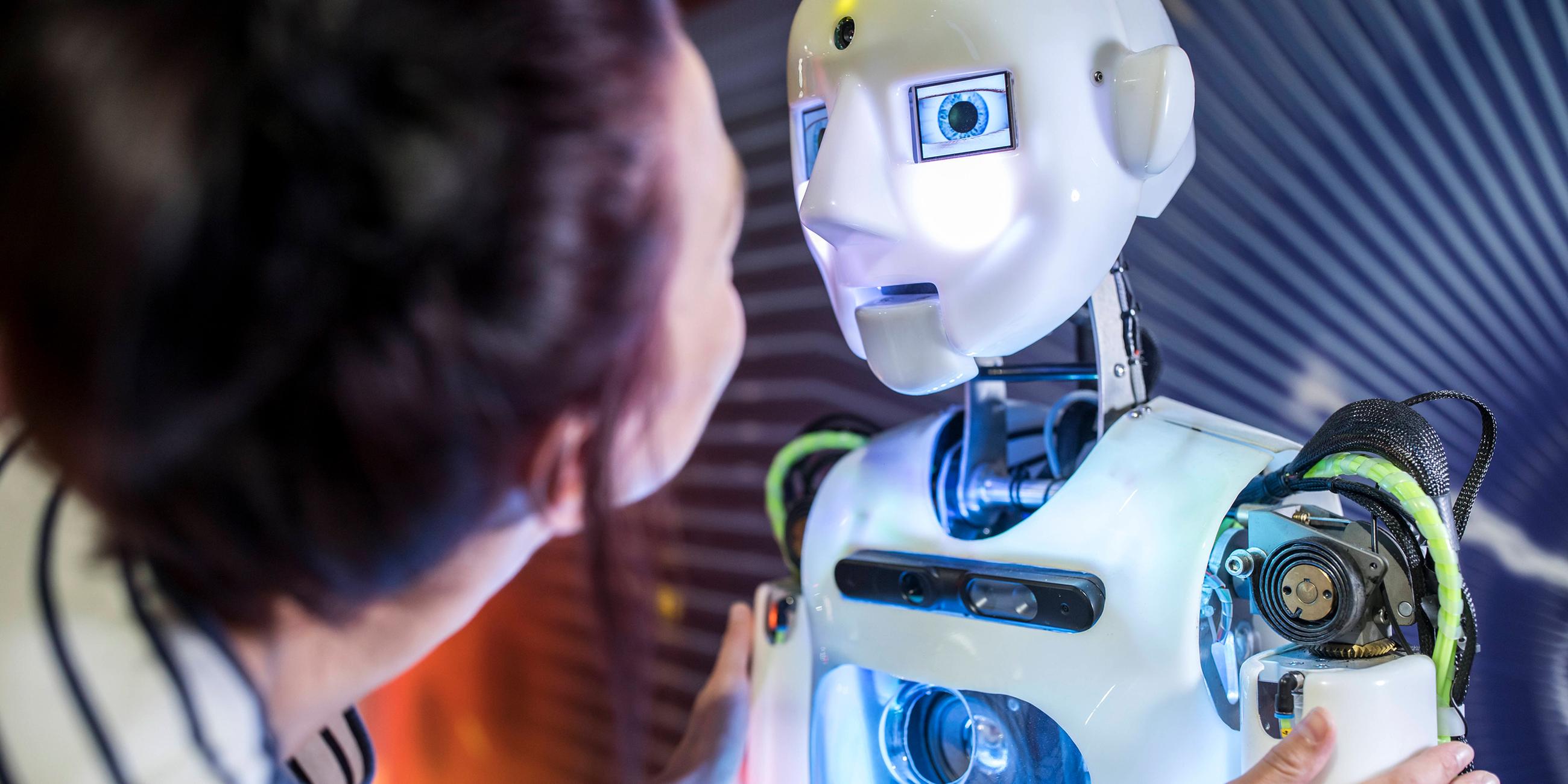 Frau beugt sich nach an einen Roboter in menschlicher Gestalt und betrachtet sein Gesicht.