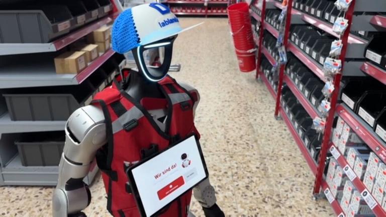 Roboter "Schotti" mit blauer Kappe und roter Weste steht im Baumarkt.