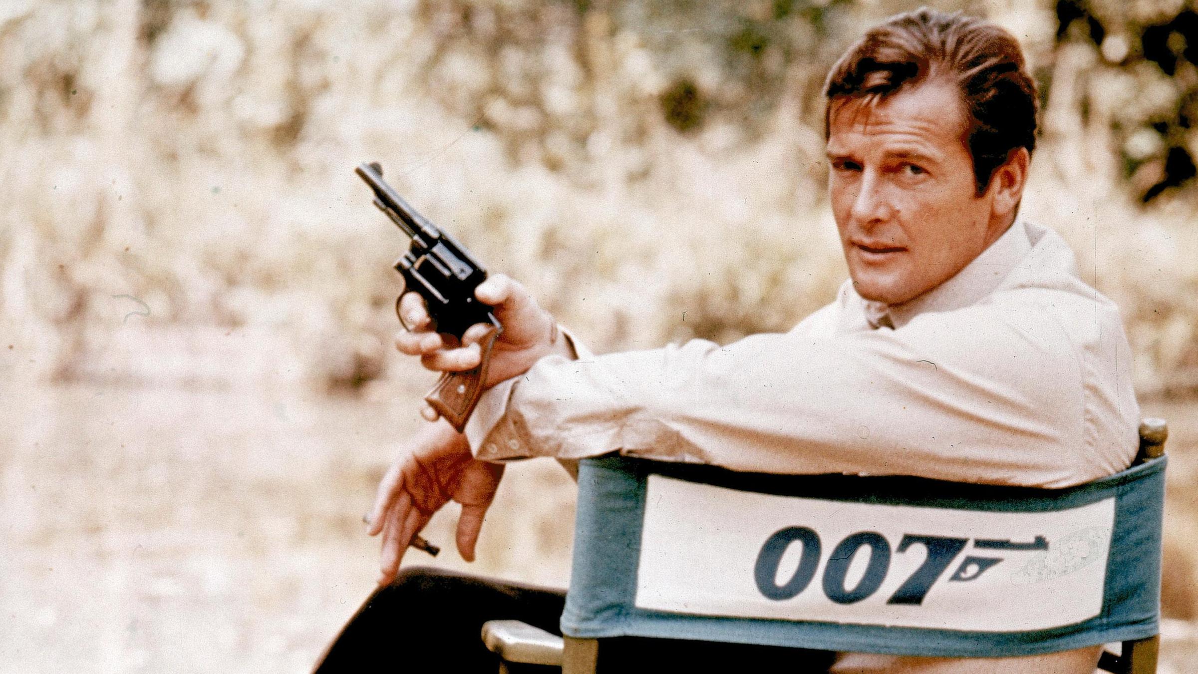 Roger Moore sitzt im Regiestuhl mit der Aufschrift 007. Er lehnt sich nach links über die Stuhllehne in die Kamera und hält eine Pistole in der rechten Hand.