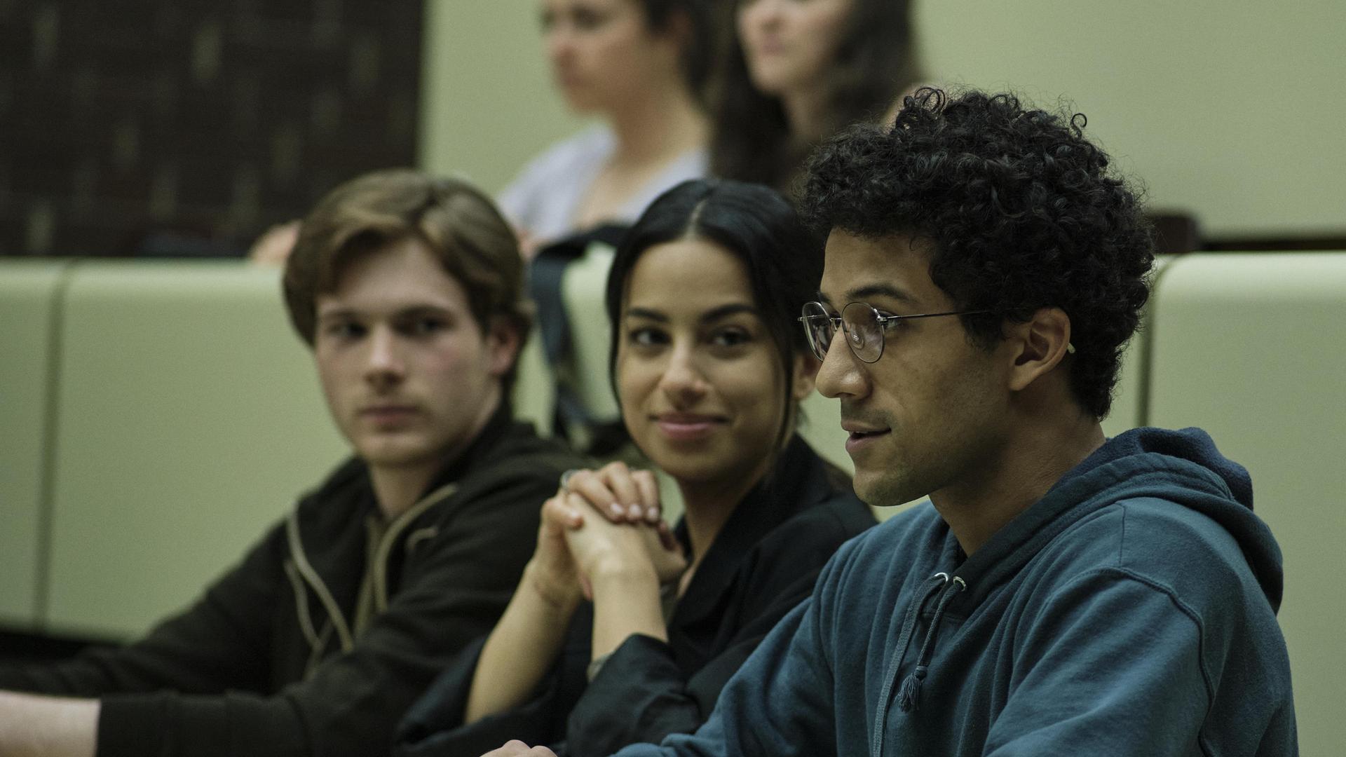 Am Ende – Die Macht der Kränkung: David, Aischa und Roko sitzen nebeneinander in einem Hörsaal. David und Aischa schauen zu Roko, dessen Blick nach vorne gerichtet ist.