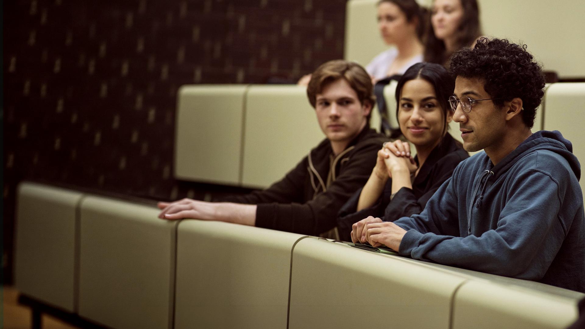Am Ende – Die Macht der Kränkung: David, Aischa und Roko sitzen nebeneinander in einem Hörsaal. David und Aischa schauen zu Roko, dessen Blick nach vorne gerichtet ist.