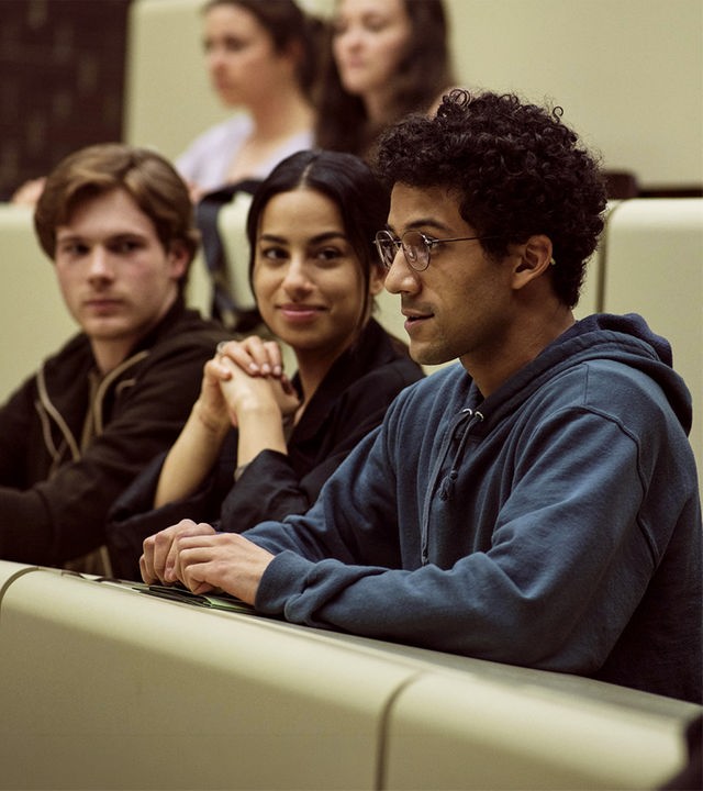 Am Ende – Die Macht der Kränkung: David, Aischa und Roko sitzen nebeneinander in einem Hörsaal. David und Aischa schauen zu Roko, dessen Blick nach vorne gerichtet ist.