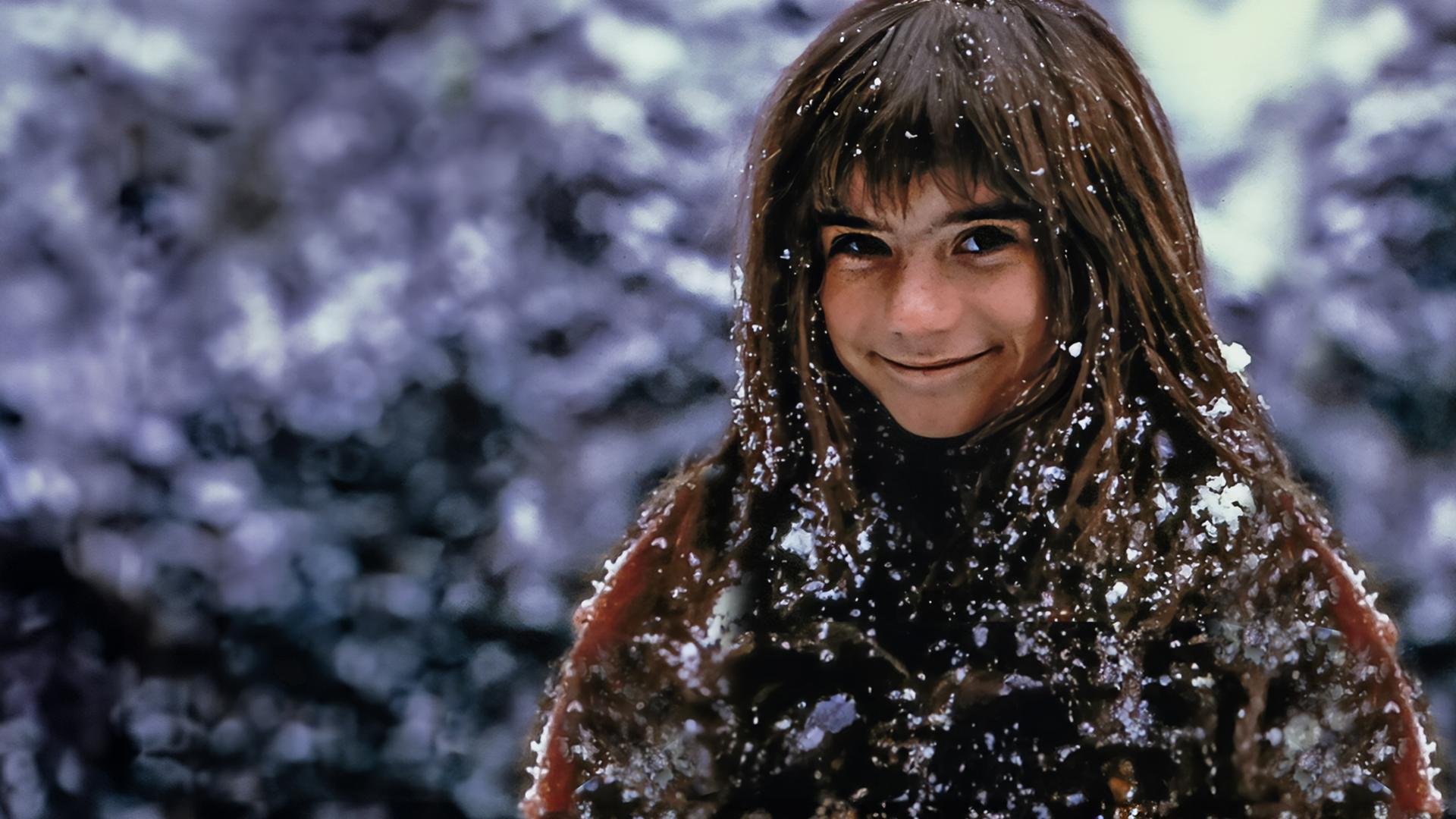 Außenaufnahme. Ronja, gespielt von Hanna Zetterberg, steht lächelnd da und ist mit Schnee bedeckt.