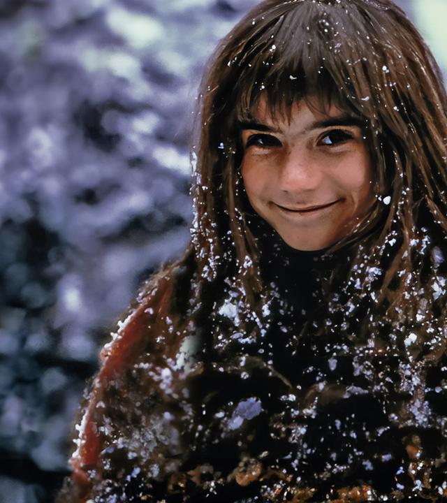 Außenaufnahme. Ronja, gespielt von Hanna Zetterberg, steht lächelnd da und ist mit Schnee bedeckt.