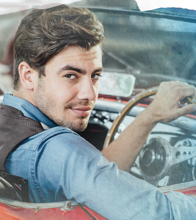 Bruno (Florian Wünsche) sitzt in seinem rot-weißen Oldtimer-Cabrio am Steuer und blickt lächelnd über die Schulter.