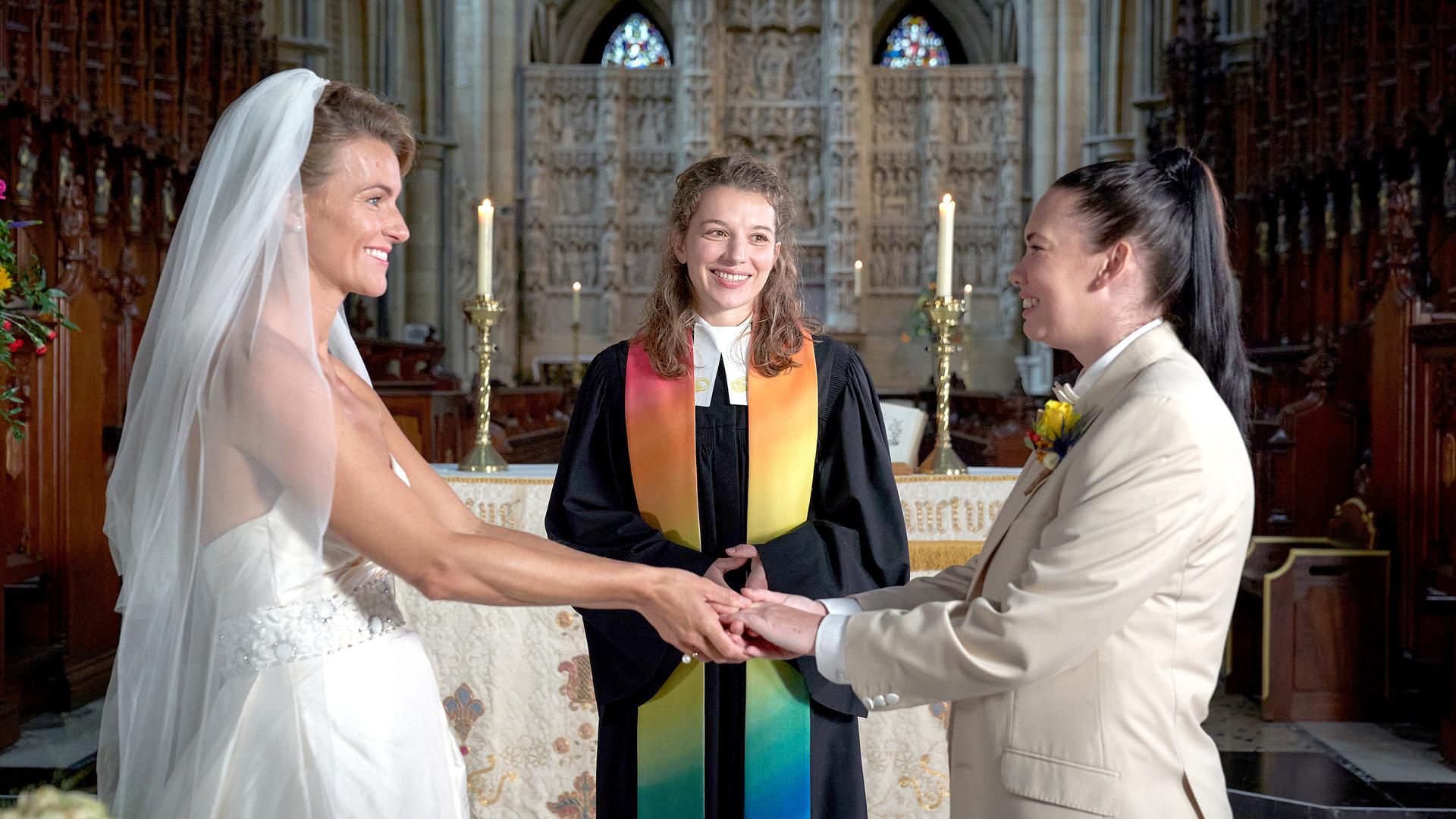 Vor dem Altar steht Vikarin Rebecca (Antonia Bill) und lächelt. Vor ihr halten sich zwei Frauen (Emma Skinner und Rachel Leigh), die eine im Brautkleid, die andere im Anzug, an den Händen und stahlen sich an.