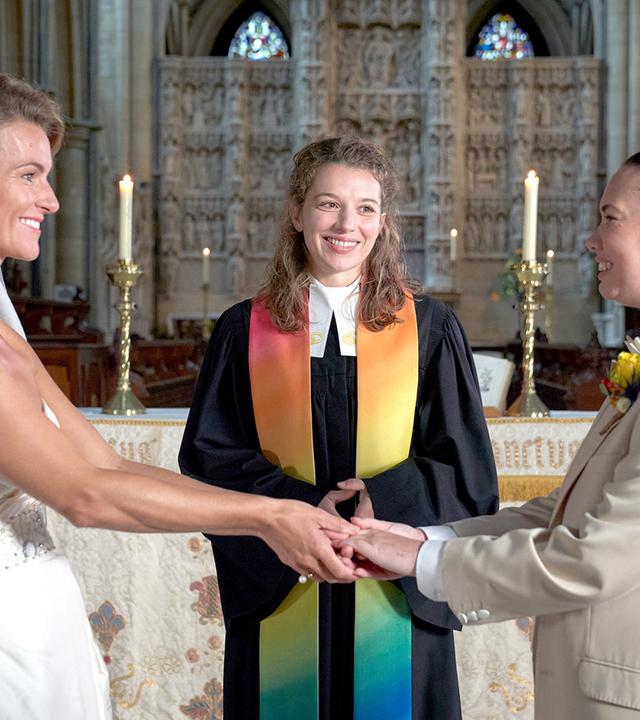 Vor dem Altar steht Vikarin Rebecca (Antonia Bill) und lächelt. Vor ihr halten sich zwei Frauen (Emma Skinner und Rachel Leigh), die eine im Brautkleid, die andere im Anzug, an den Händen und stahlen sich an.