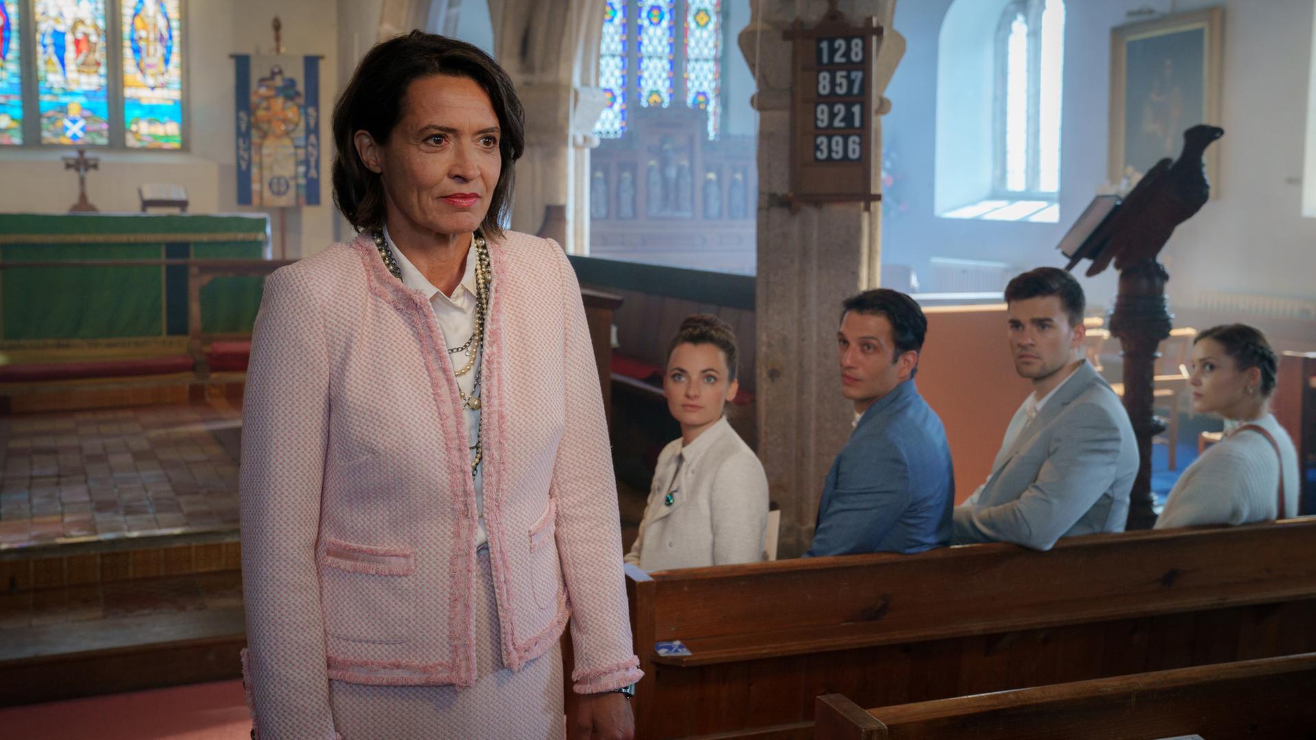 "Rosamunde Pilcher: Schwiegertöchter": In vorderstet Kirchenbank sitzen Kim (Paula Schramm), Patrick (Max Hemmersdorfer), Robin (Patrick Mölleken) und Marisa (Maja Céline Probst) und blicken erwartungsvoll auf Lynette (Ulrike Folkerts). Diese steht steht zwischen den Bänken und lächelt.