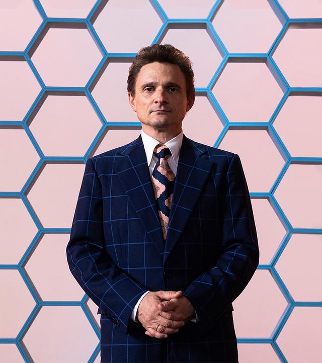 Hans Rosenthal blickt mit einem leichten Lächeln und vor dem Körper zusammengefalteten Händen in die Kamera. Er trägt einen dunkelblauen Anzug mit grobkariertem Muster.