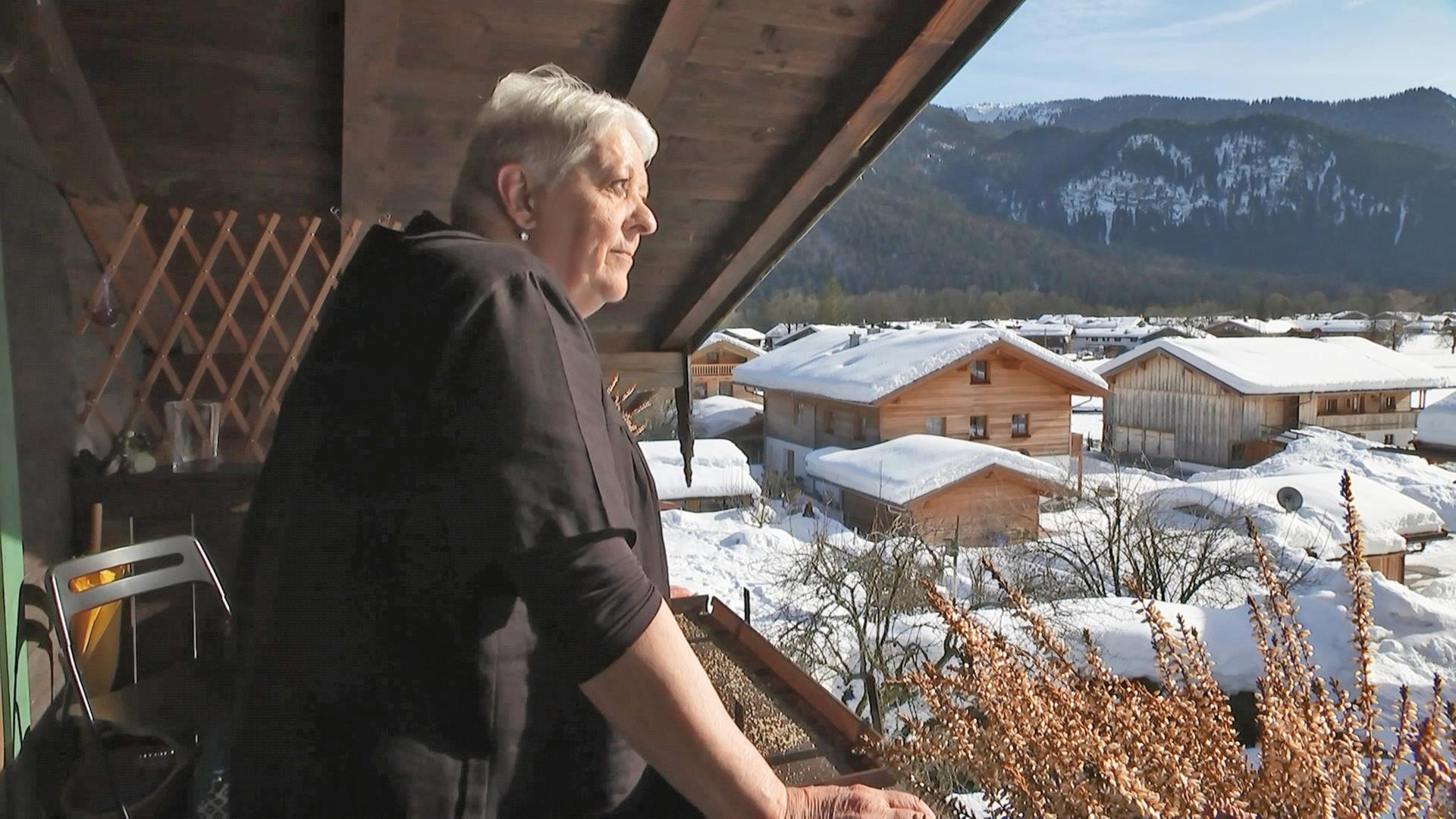 Eine weißhaarige Frau steht auf dem Balkon einen Holzhauses und schaut in die verschneite Gegend.