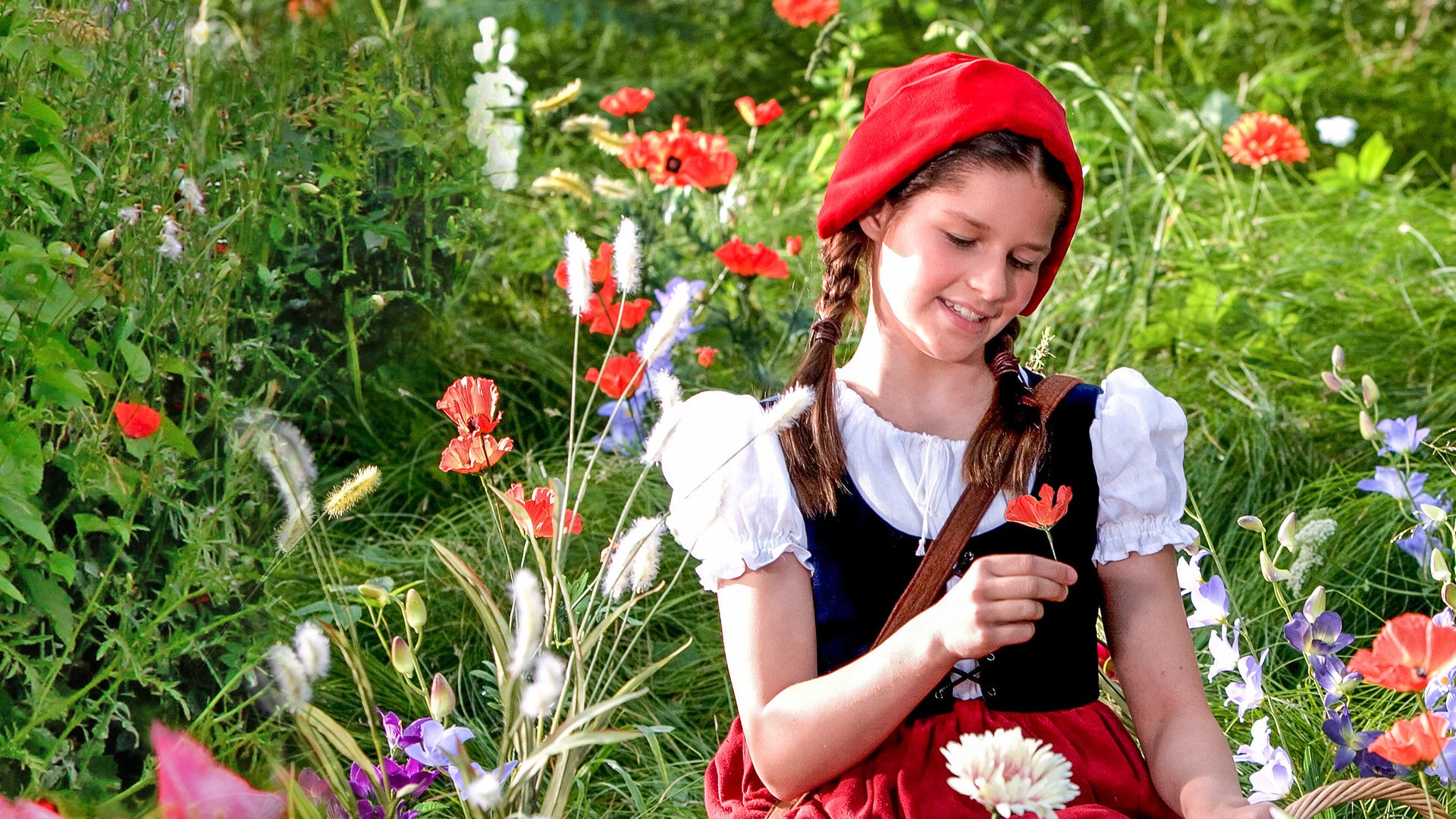 Rotkäppchen (Kathleen Frontzek) sitzt auf einer Blumenwiese. In der rechten Hand hält sie eine Mohnblume, mit der linken Hand stützt sie sich auf ihrem Körbchen ab. Sie trägt einen roten Rock, eine weiße Bluse und eine kleine schwarze Weste. Auf dem Kopf hat sie eine rote Kappe.