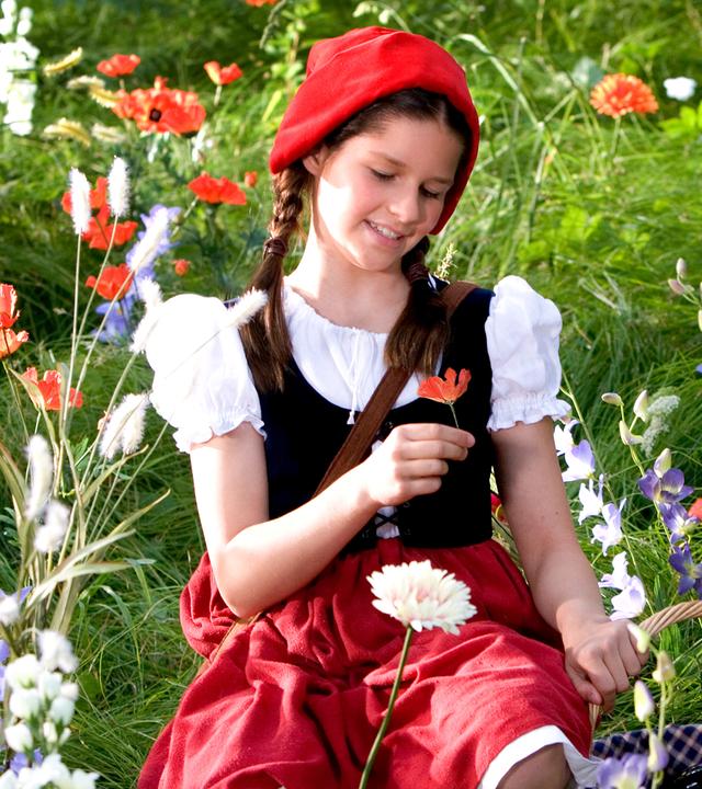 Rotkäppchen (Kathleen Frontzek) sitzt auf einer Blumenwiese. In der rechten Hand hält sie eine Mohnblume, mit der linken Hand stützt sie sich auf ihrem Körbchen ab. Sie trägt einen roten Rock, eine weiße Bluse und eine kleine schwarze Weste. Auf dem Kopf hat sie eine rote Kappe.