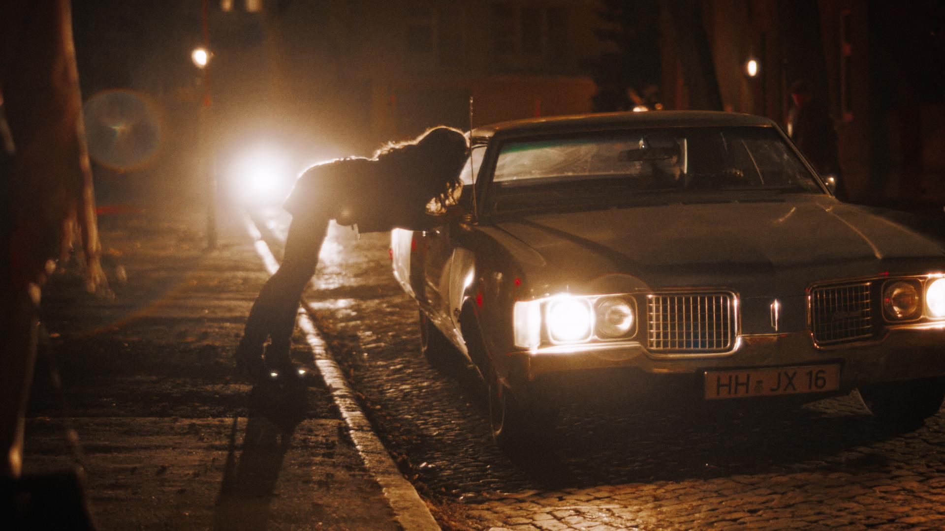 Ein Auto hält nachts am Straßenrand. Die Scheinwerfer leuchten, während sich eine junge Frau zum Beifahrerfenster herein beugt, um mit dem Fahrer zu sprechen.